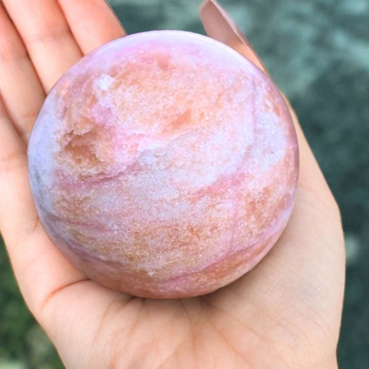 High grade polished pink opal sphere showing rich pastel pink tones and natural cream banding held in hand.