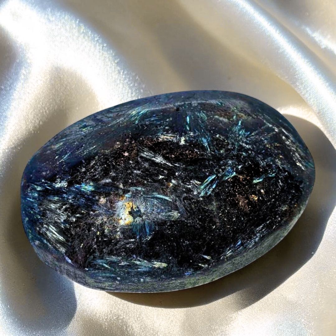 A polished dark blue and black fireworks crystal stone with shimmering blue and silver needle blades on satin